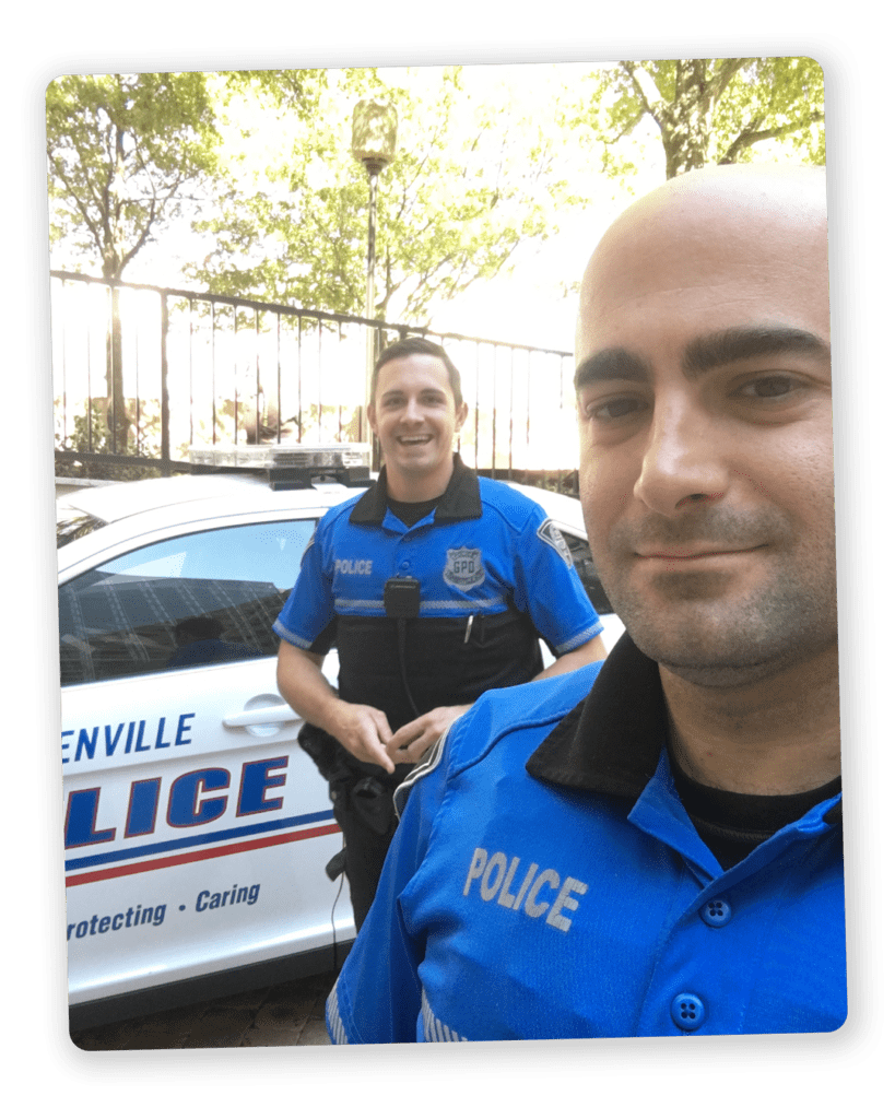 Two law enforcement officers (LEO) smiling at the camera in the street with a Greenville police car in the background
